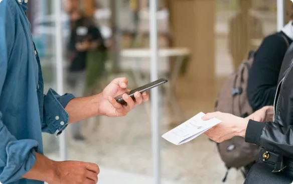 Persona entregando una entrada impresa a otra persona mientras sostiene un smartphone, ilustrando el uso de impresoras de tarjetas y entradas térmicos.