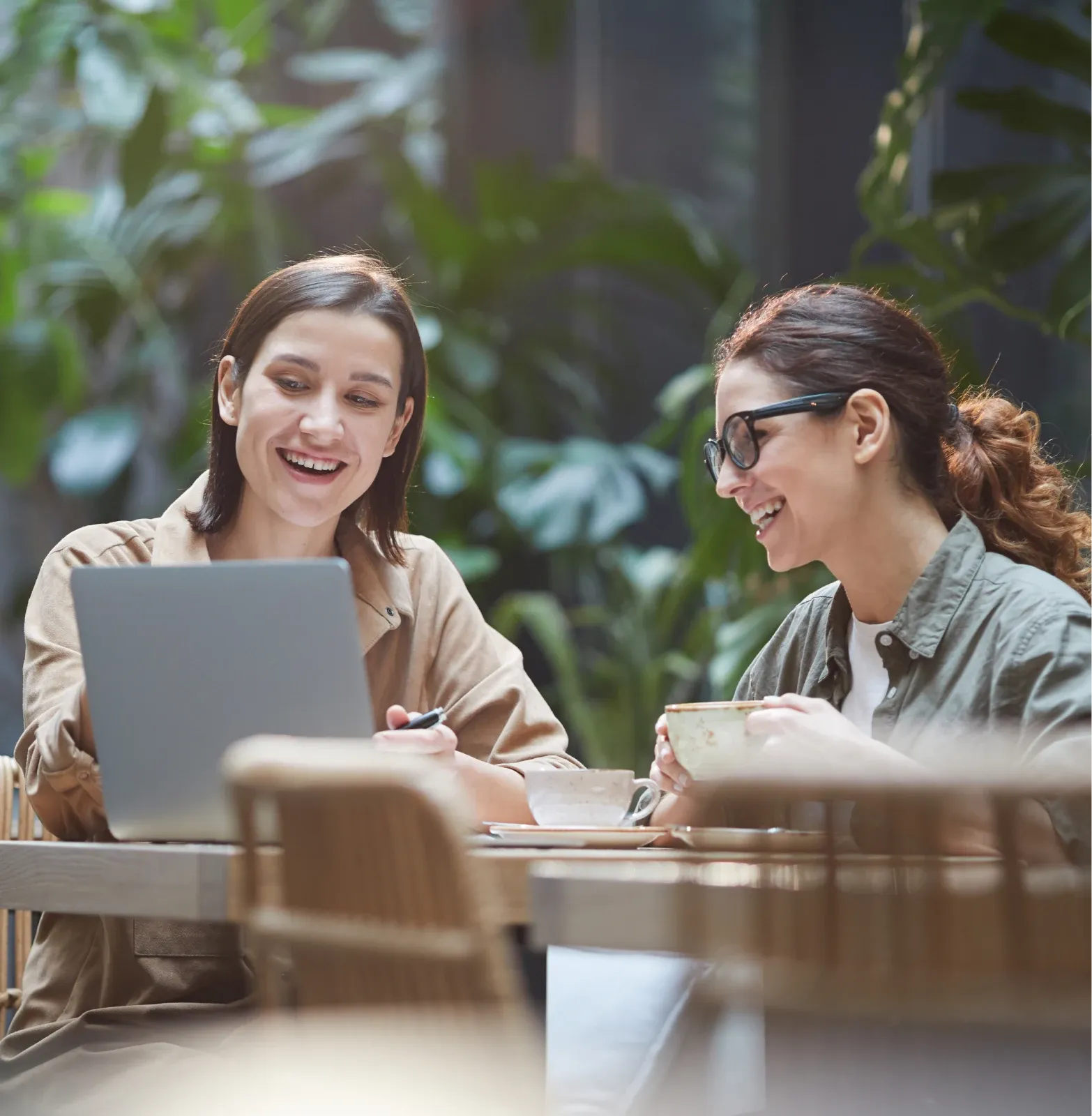 Dos mujeres sonrientes trabajan juntas en un portátil, sentadas a una mesa en un espacio luminoso y frondoso rodeado de plantas.